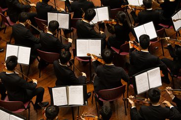 An image showing the strings section of an orchestra from above, with musicians seated and playing on violins.